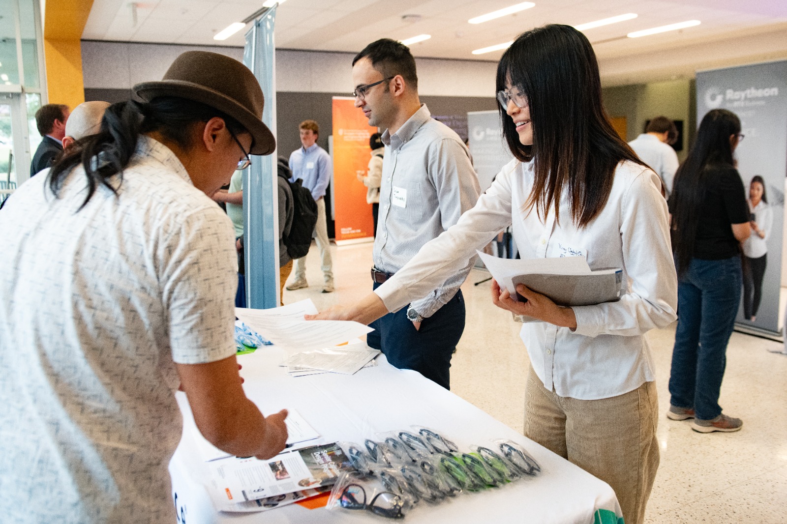 Person hands out materials at a table during the ASE/EM Industry Open House 2024, with attendees engaging in conversations near company booths in the background.