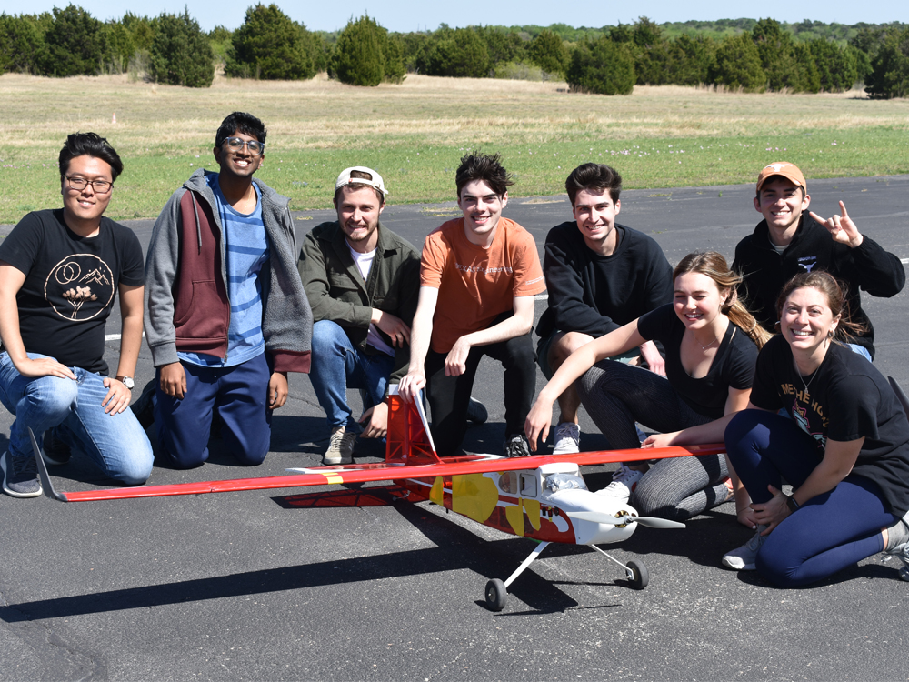 Group of students smiling with aircraft prototype in field.