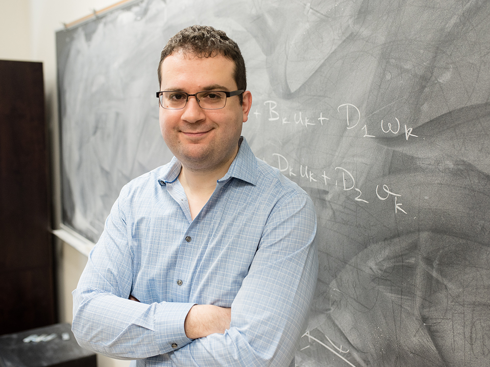 Efstathios Bakolas smiling in front of chalkboard with arms crossed.