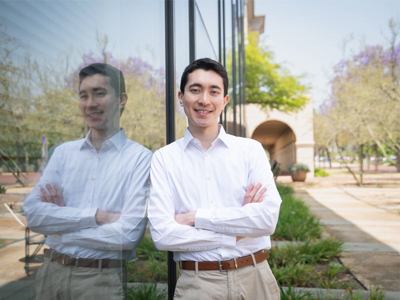 Nicholas Nelsen smiling and leaning against glass window outside with arms crossed in front of him.