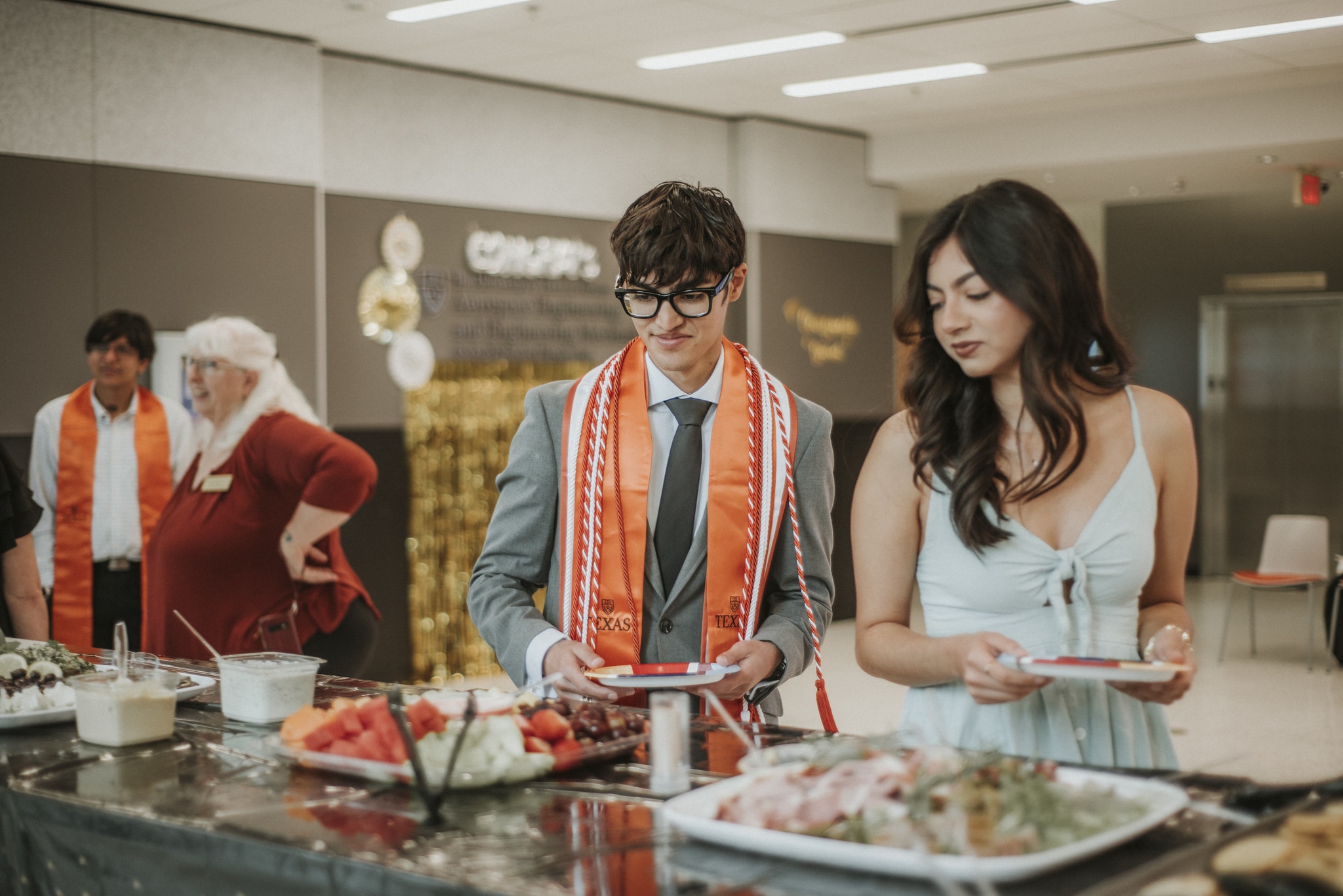 Students wearing graduation regalia filling plates with food at a buffet table.