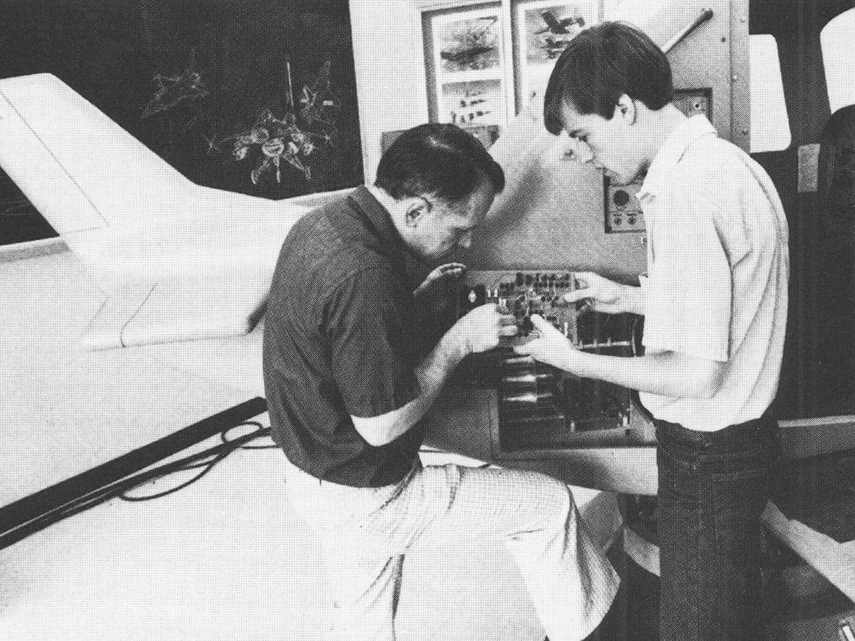 Two people inspect and work on an exposed electronic system inside an aerospace laboratory.