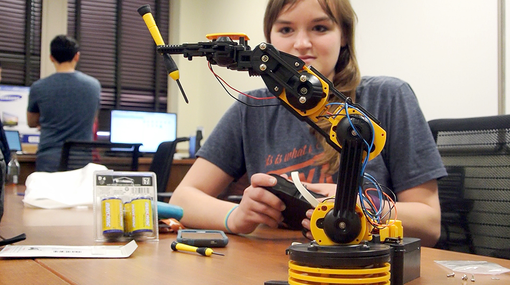 Student holding control device for robot arm being tested on table.