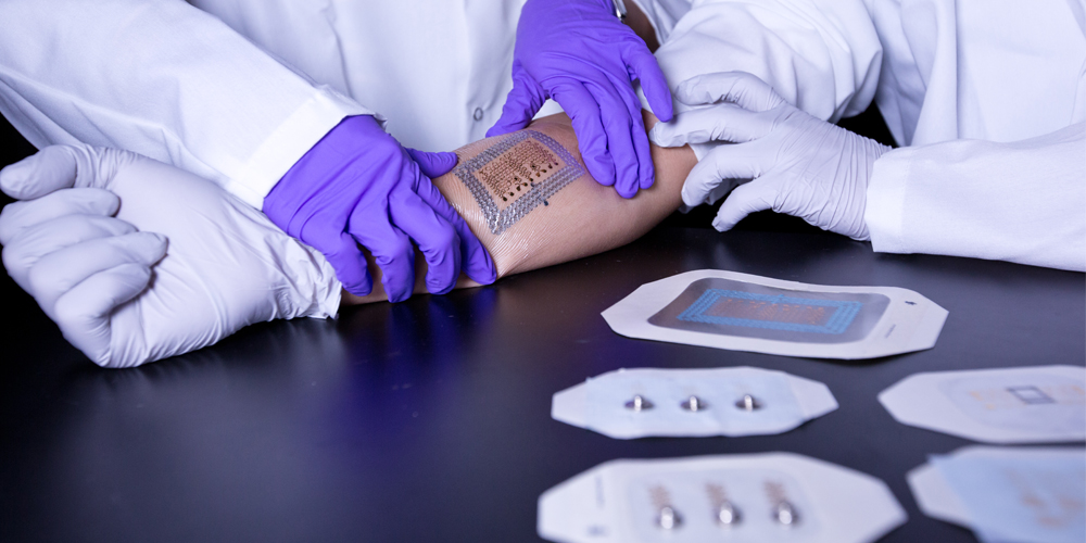 Gloved hands of a researcher applying an electronic tattoo prototype to another scientist's bare arm.