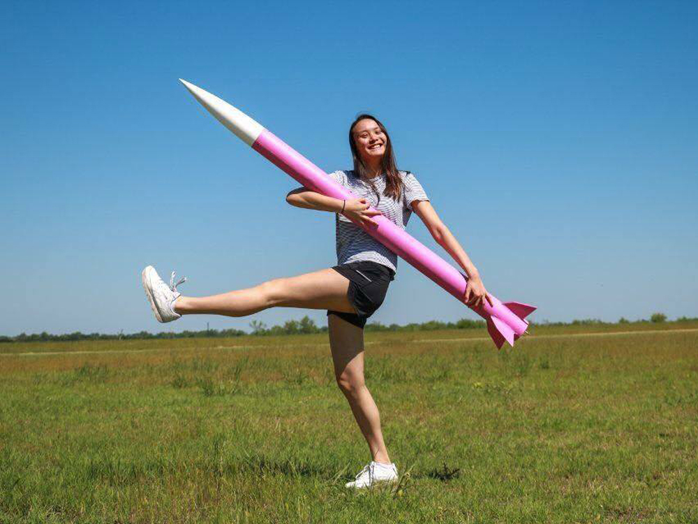 Anna Wei holding rocket model and kicking her leg upwards in a field.